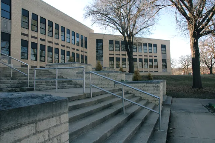 Brick school buildings seen across concrete steps, handrails and a tree-lined lawn.
