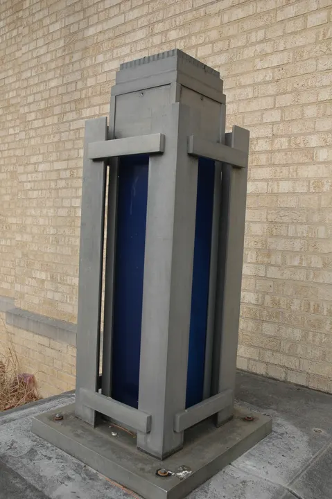 A blue-and-metal geometric fixture on a base beside a brick wall.